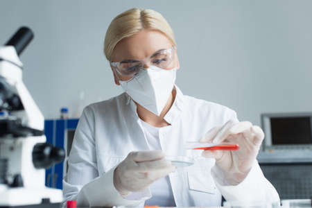 Scientist in goggles and latex gloves holding test tube and petri dish near blurred microscope in...