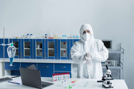 Scientist in hazmat suit and latex gloves standing near test tubes and microscope in labの写真素材