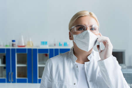 Scientist in goggles and protective mask talking on smartphone in labの写真素材