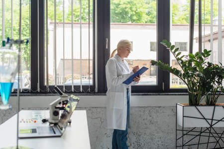 Side view of scientist writing on clipboard near window in laboratory