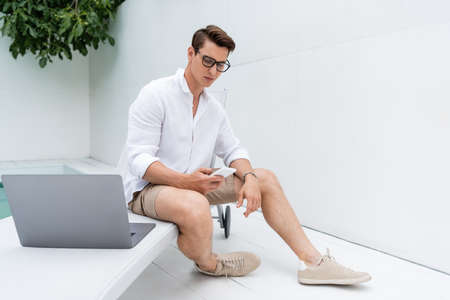 full length of man in sneakers sitting at poolside near laptop and messaging on mobile phoneの写真素材