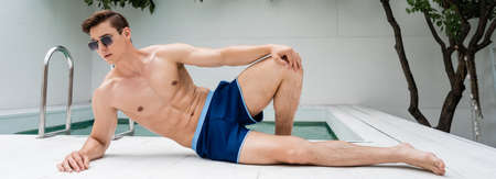 full length of athletic man in swimming trunks and sunglasses resting at poolside, bannerの写真素材