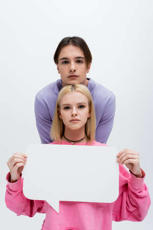 Brunette man looking at camera near girlfriend with white speech bubble isolated on grey