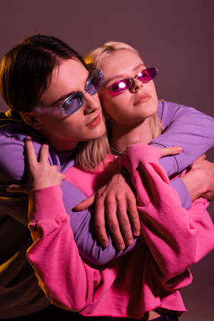 Stylish man in sunglasses embracing girlfriend isolated on purple with lightingの写真素材