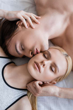Top view of shirtless man kissing blonde girlfriend with closed eyes on bed