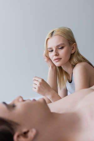 Blonde woman looking a blurred shirtless boyfriend on bed isolated on grey