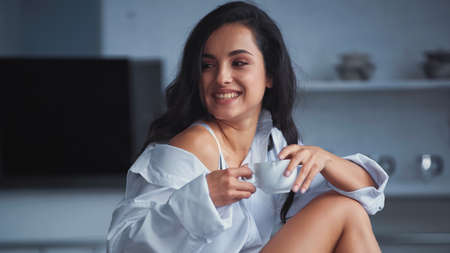 happy young woman in white shirt smiling while holding cup of coffee in morning