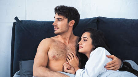 shirtless boyfriend and smiling girlfriend in white shirt looking away while lying on bed