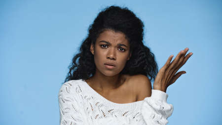 displeased african american woman in white knitted sweater gesturing isolated on blue