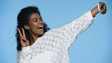 happy african american woman taking selfie on smartphone and showing peace sign isolated on blueの写真素材
