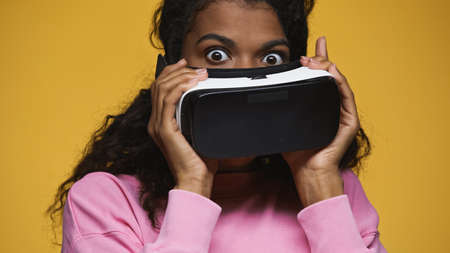 surprised african american woman in pink sweatshirt holding vr headset isolated on yellow