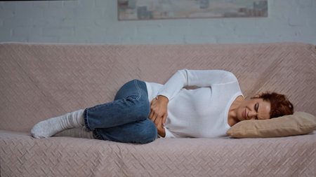 young woman lying on sofa and suffering from cramps and having stomach ache