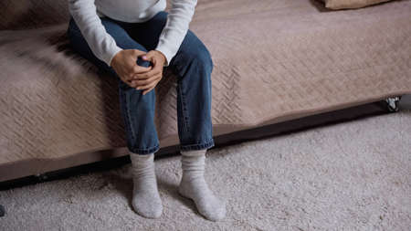cropped view of woman in jeans feeling knee pain while sitting on sofa in living room
