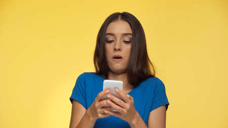 young brunette woman in blue t-shirt grimacing while typing on smartphone isolated on yellow