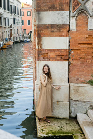 Pregnant woman in dress holding string bag near river and building in Veniceの写真素材
