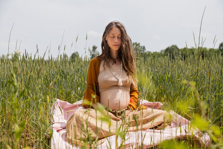 Pregnant woman with closed eyes sitting on blanket in fieldの写真素材
