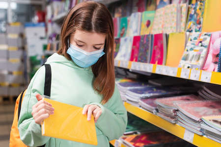 child in medical mask holding notebook while choosing school supplies in shop