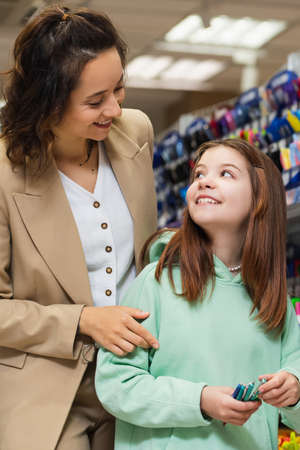 cheerful woman looking at happy daughter holding new pens in stationery shop