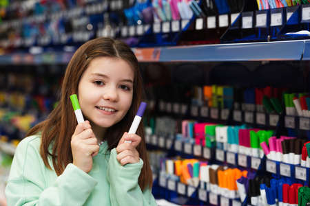 smiling schoolgirl holding colorful felt pens and looking at camera in stationery store