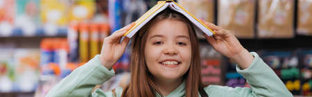 cheerful girl covering head with copybook and looking at camera in blurred stationery shop, banner