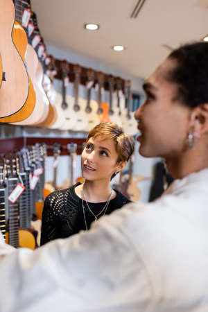 Smiling customer looking at acoustic guitars near blurred african american seller in music storeの写真素材