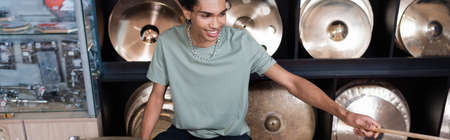 Cheerful african american customer holding drumstick near plates in music store, banner