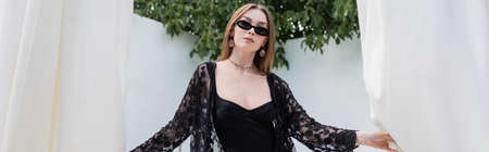 Stylish woman in swimsuit and sunglasses holding curtains at resort, banner
