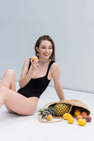 Cheerful young woman in swimsuit holding peach near fruits in straw handbag at resortの写真素材