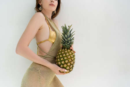 cropped view of woman in golden swimwear and dress holding ripe pineapple near while wallの写真素材