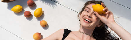 top view of happy young woman in black swimsuit lying near fresh fruits on white surface, banner