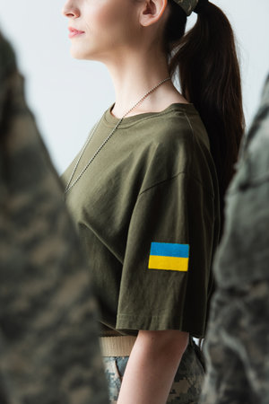 Cropped view of servicewoman with ukrainian flag on chevron standing near blurred soldiers isolated on greyの写真素材