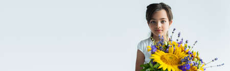 Girl looking at camera near blue and yellow flowers isolated on grey, banner