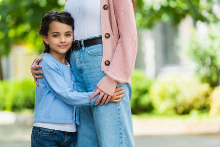 smiling girl hugging mother and looking at camera outdoors