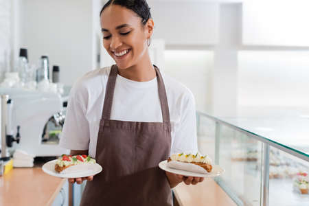 Smiling african american saleswoman holding eclairs in sweet shopの写真素材