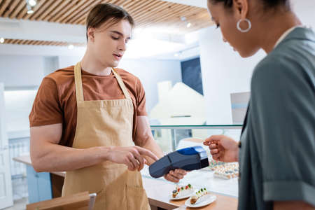 Salesman holding payment terminal near blurred african american customer in sweet shop