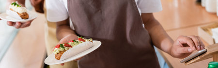 Cropped view of african american saleswoman holding dessert and using digital tablet in sweet shop,...