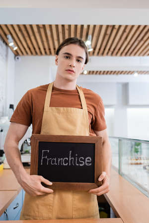 Young seller in apron holding chalkboard with franchise lettering in confectioneryの写真素材
