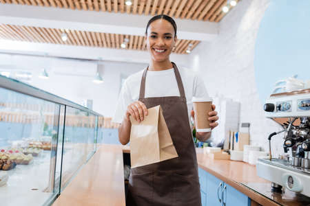 Positive african american saleswoman holding takeaway drink and paper bag in confectioneryの写真素材