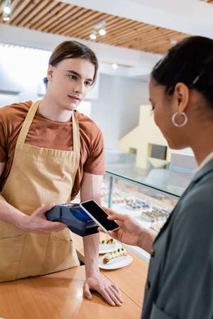 Young salesman holding payment terminal near african american customer with smartphone in confectioneryの写真素材