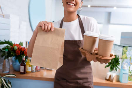 Cropped view of african american seller holding paper bag and takeaway coffee in confectioneryの写真素材