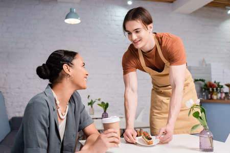 Cheerful seller giving dessert to african american customer with coffee to go in confectioneryの写真素材