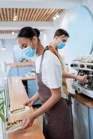 African american seller in medical mask holding eclairs near showcase in confectioneryの写真素材