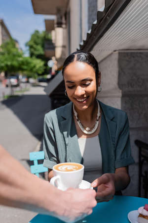 Smiling african american woman taking cappuccino from waiter near dessert on terrace of sweet shopの写真素材