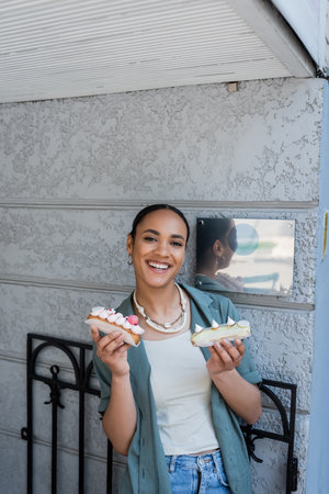 Cheerful african american woman holding sweet eclairs near confectionery