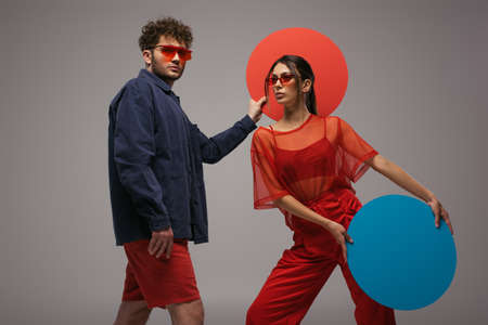 young stylish couple in blue and red outfits holding round shape carton isolated on grey