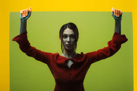 stylish young woman in red dress looking at camera through glass isolated on greenの写真素材
