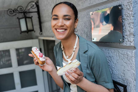 Positive african american customer holding sweet eclairs near confectionery outdoors