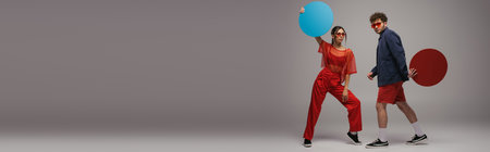 full length of young couple in trendy outfits and sunglasses holding round shape carton on grey, bannerの写真素材