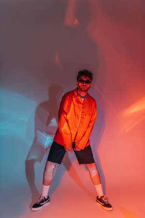 full length of curly and stylish man in sunglasses posing in studio with colorful lightの写真素材
