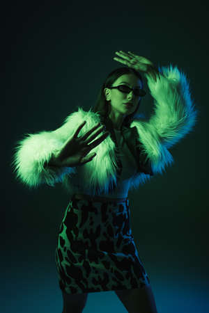 stylish young woman in faux fur jacket and sunglasses gesturing while posing on blueの写真素材